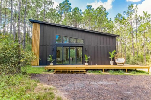 Modern black house with wooden accents, surrounded by trees, featuring a deck and large glass windows.