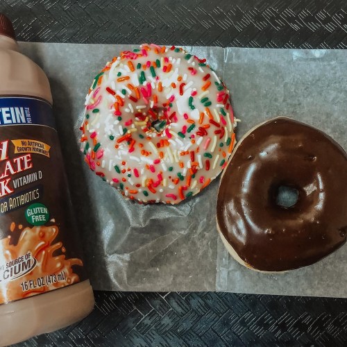 A chocolate donut and a frosted donut with sprinkles, alongside a bottle of chocolate protein drink on parchment paper.