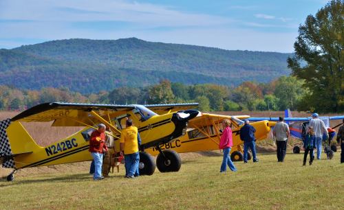 Two yellow planes parked on a grassy field with people gathered around, set against a backdrop of mountains and blue sky.
