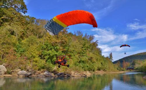 Two powered paragliders soar above a river surrounded by lush greenery and rocky banks under a clear blue sky.