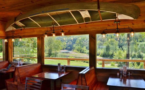 Cozy restaurant interior with wooden booths, a canoe overhead, and large windows showcasing a scenic view of greenery.
