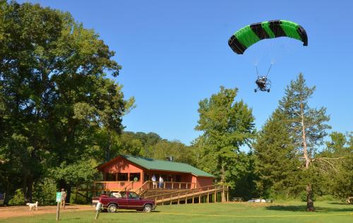 A powered paraglider flies above a cabin surrounded by trees, with people on the porch and a truck parked nearby.