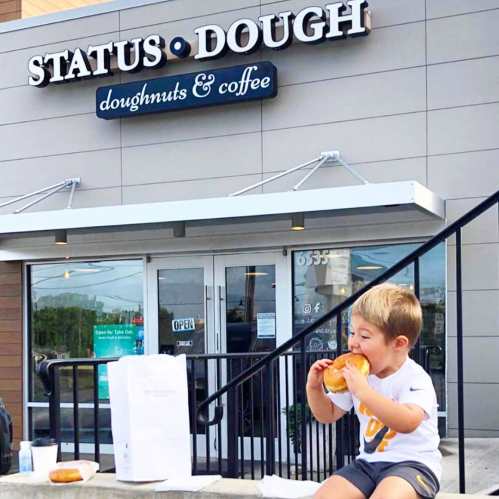 A young child sits on stairs, happily eating a doughnut outside a doughnut shop named "Status Dough."