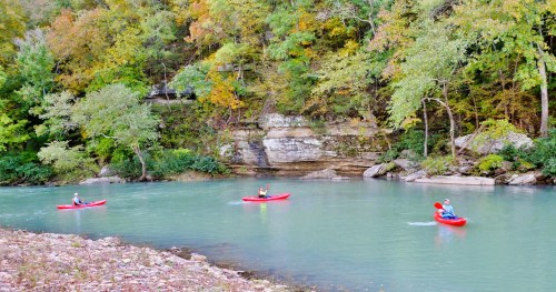 Three kayakers in red kayaks paddle on a calm, turquoise river surrounded by autumn-colored trees and rocky cliffs.