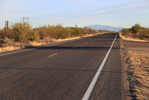 A long, empty road stretches through a desert landscape with sparse vegetation and distant mountains under a clear sky.