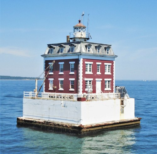 A red and white lighthouse stands on a rocky base in the ocean, surrounded by calm blue waters under a clear sky.