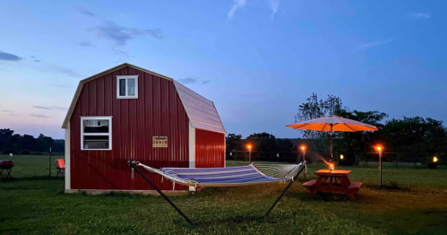 A cozy red barn at dusk, featuring a hammock, picnic table, and an umbrella, surrounded by a grassy field.