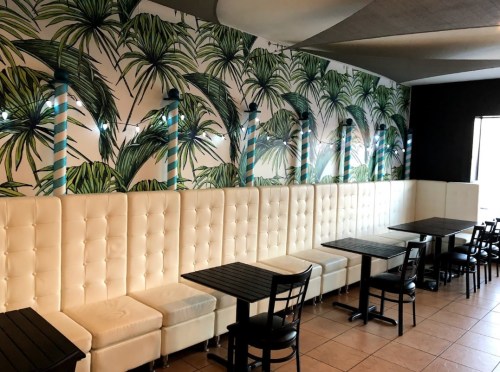 A modern cafe interior with palm leaf wallpaper, white tufted seating, and black tables and chairs.