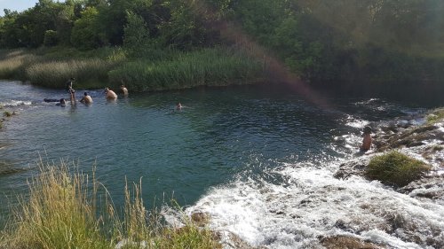 People swimming and playing in a natural water body surrounded by greenery and a small waterfall.
