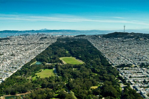 Aerial view of a cityscape divided by a large green park, with water and hills in the background.