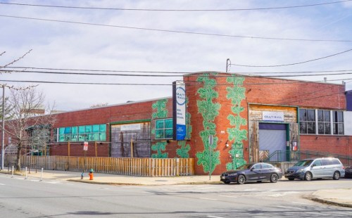 A brick building with green leaf murals, surrounded by a wooden fence and parked cars on a street.