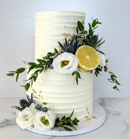 A three-tiered white cake adorned with fresh flowers, greenery, and a lemon slice, set on a marble surface.