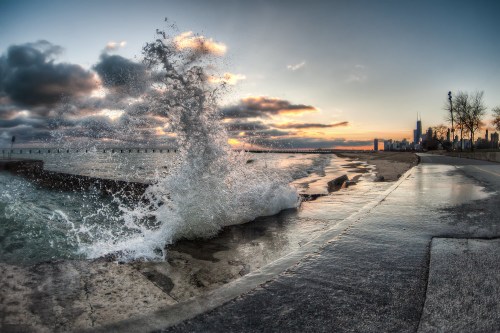 A wave crashes against a shoreline at sunset, with a city skyline in the background and dramatic clouds overhead.