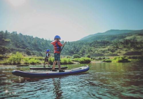 Two children paddleboarding on a calm river surrounded by lush greenery and mountains under a clear blue sky.