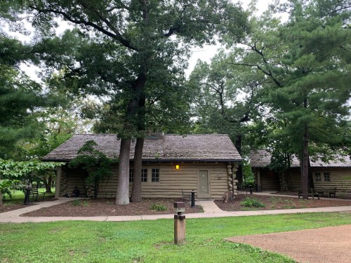 A rustic log cabin surrounded by trees and greenery, with a path leading to the entrance.