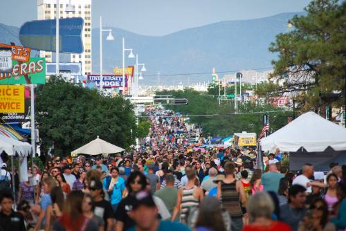 A bustling street filled with crowds at a festival, with tents and mountains in the background.