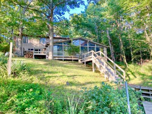 A wooden house with large windows, surrounded by trees, set on a grassy slope with a staircase leading up to it.