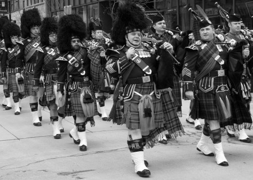 A group of bagpipers in traditional Scottish attire marching in a parade, wearing kilts and feathered hats.