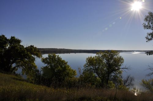 A serene lake reflects the clear blue sky, framed by lush green trees and sunlight glinting off the water's surface.