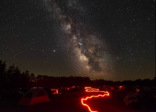 A starry night sky with the Milky Way above a campsite, featuring glowing red trails on the ground.