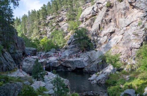 A scenic rocky landscape with a swimming hole, surrounded by trees and people enjoying the outdoors.