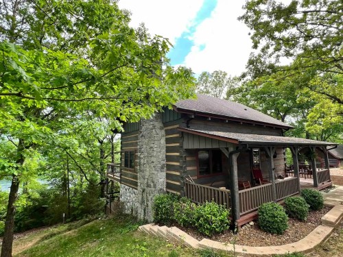 A rustic cabin surrounded by trees, featuring a porch and stone accents, near a serene body of water.