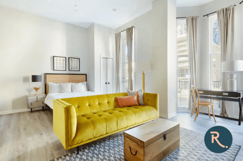 Bright, modern bedroom with a yellow sofa, wooden coffee table, and a desk by a large window.
