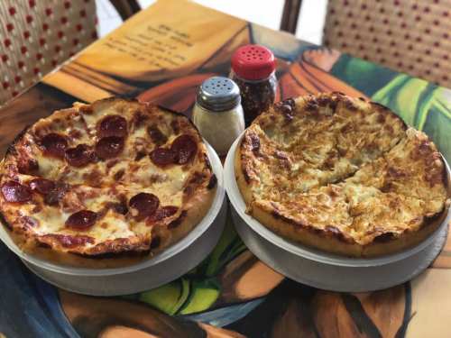 Two pizzas on a table: one with pepperoni and cheese, the other with a white sauce and toppings, alongside condiments.