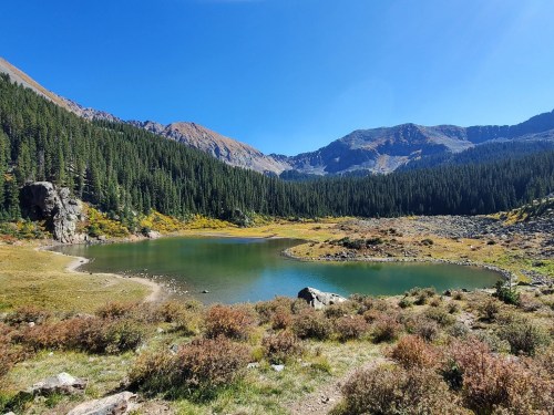 A serene mountain lake surrounded by lush green trees and rocky terrain under a clear blue sky.