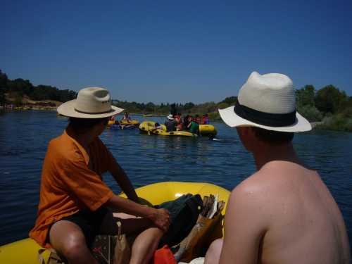 Two people in hats sit in a yellow raft, watching others in kayaks on a sunny river. Green trees line the shore.