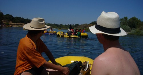 Two people in hats sit in a yellow raft, watching others float down a river on a sunny day.