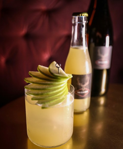 A refreshing cocktail garnished with apple slices, with a bottle and a sparkling drink in the background.
