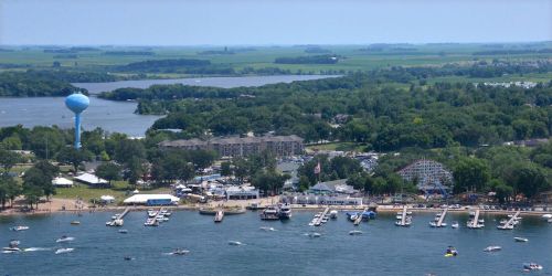 Aerial view of a lakeside area with boats, a beach, and a water tower in the background surrounded by greenery.