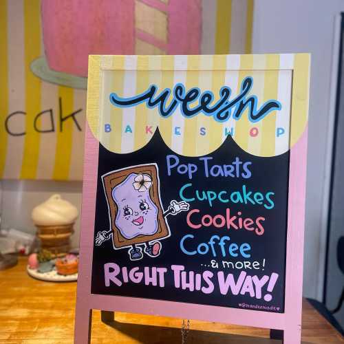 Colorful sign for Weezh Bakeshop featuring Pop Tarts, cupcakes, cookies, coffee, and an arrow pointing right.