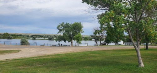 A serene landscape featuring a calm river, green trees, and a grassy area under a cloudy sky.
