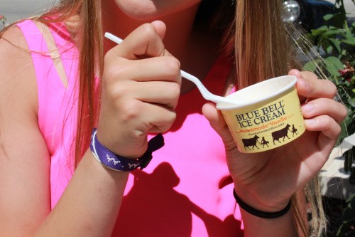 A person in a pink shirt holds a small cup of Blue Bell vanilla ice cream with a spoon.