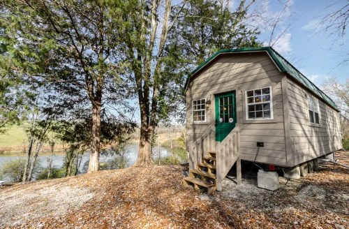 A small, cozy cabin with a green door, surrounded by trees and near a calm river, set in a scenic outdoor landscape.