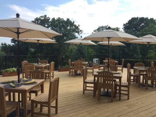 Outdoor dining area with wooden tables and umbrellas, surrounded by greenery under a partly cloudy sky.