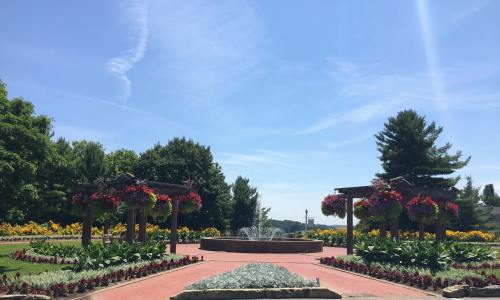 A sunny park scene featuring flower beds, a fountain, and decorative pergolas with hanging plants.