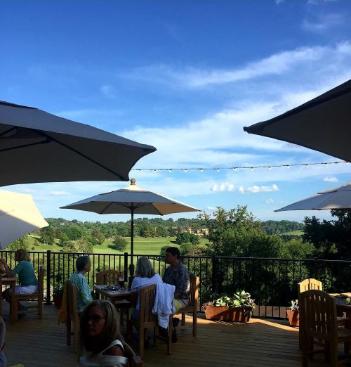 Outdoor dining area with umbrellas, overlooking a green landscape and blue sky, with people enjoying their meals.