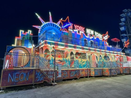 A brightly lit carnival ride themed like New York City, featuring neon lights and iconic landmarks at night.
