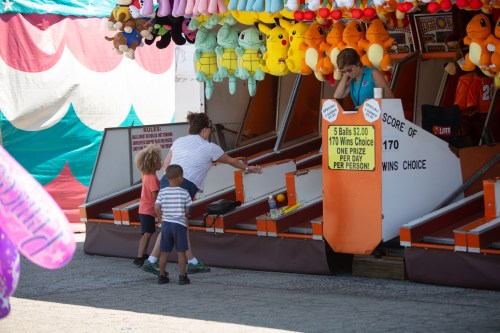 A carnival game booth with colorful prizes, where children play while an attendant watches. Bright, festive atmosphere.