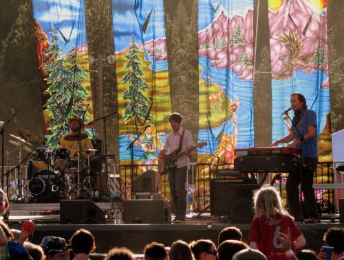 A band performs on stage with colorful backdrop art, while an audience enjoys the music in the foreground.