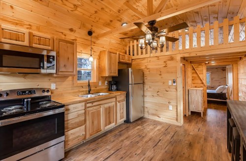 Cozy wooden cabin interior featuring a kitchen, dining area, and loft with a ceiling fan and warm wood finishes.