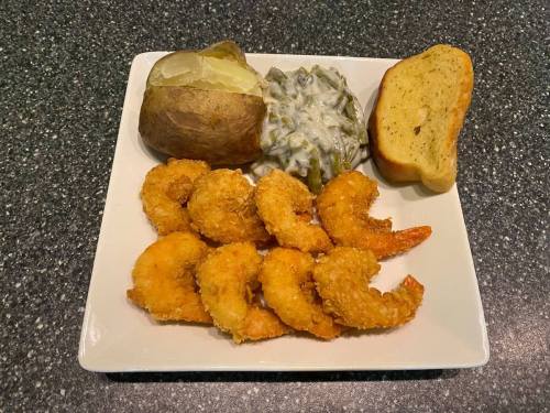 A plate with fried shrimp, a baked potato, green beans, and a slice of garlic bread on a dark countertop.