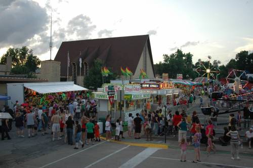 A lively fair scene with colorful stalls, crowds of people, and a ferris wheel in the background under a cloudy sky.