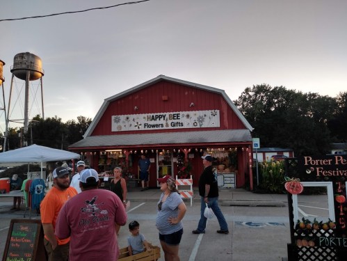 A red building with "Happy Bee Flowers & Gifts" sign, people walking outside, and a water tower in the background.