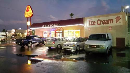 A donut shop and ice cream parlor with parked cars outside, reflecting in wet pavement under a cloudy sky.