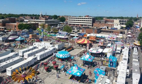 Aerial view of a vibrant fairground with colorful rides, tents, and crowds enjoying a sunny day.