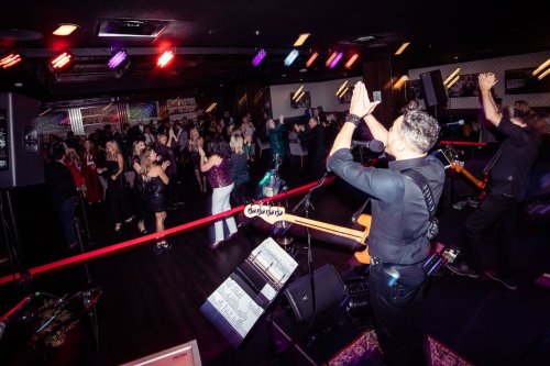 A lively crowd dances at a venue while a band performs on stage, with colorful lights illuminating the scene.
